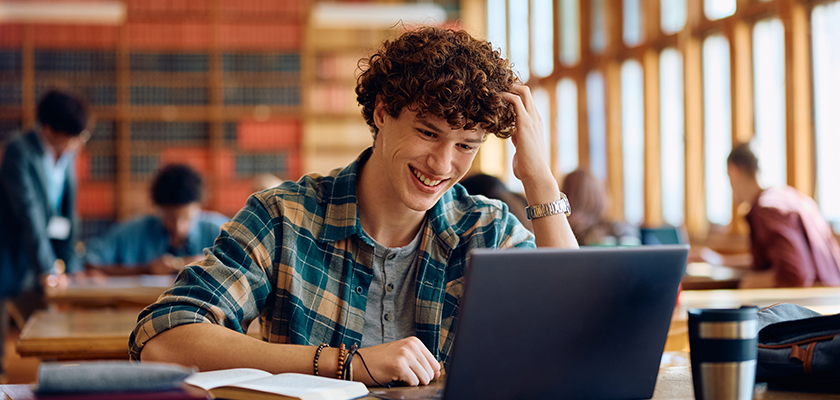 Male student doing schoolwork on a laptop in the library while smiling